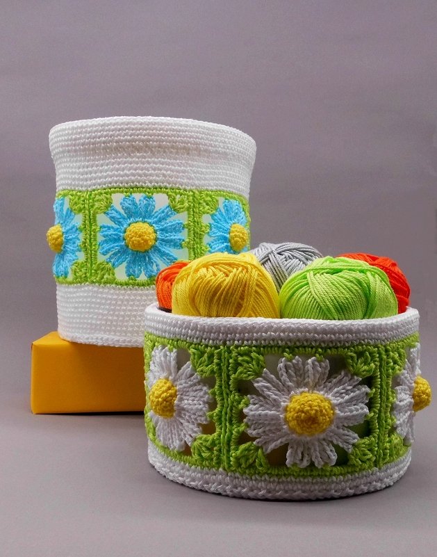 Zwei gehäkelte Körbe mit Gänseblümchen‑Motiv, einer gefüllt mit bunten Garnknäueln.