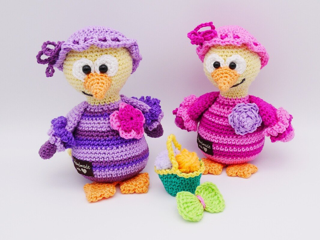 Zwei gehäkelte Amigurumi-Vögel in Rosa und Lila mit Mützchen, Blumenapplikationen, kleinem Korb und grüner Schleife