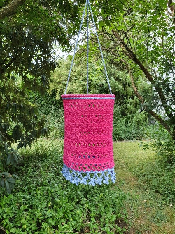 Pinker gehäkelter Hängekorb mit blauem Rand an einem Ast im Garten