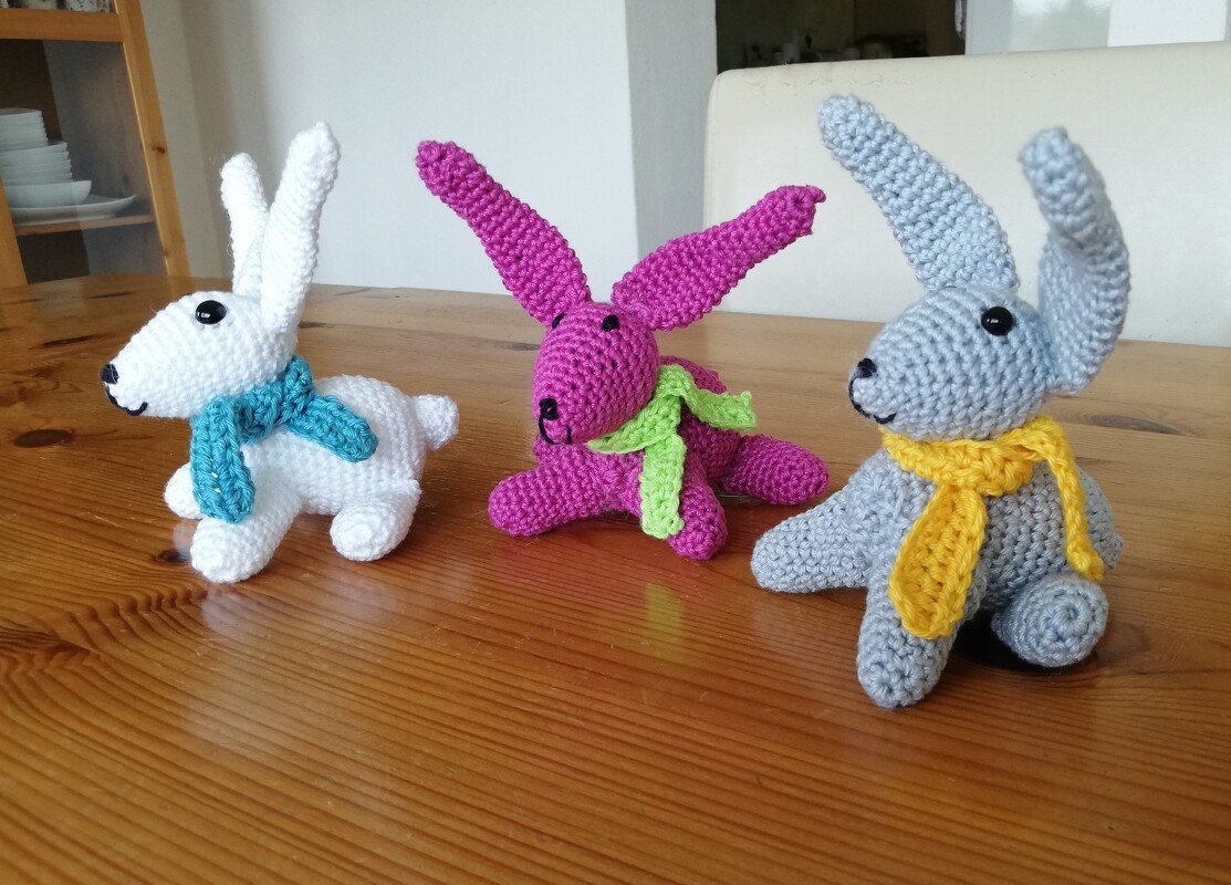 Drei gehäkelte Amigurumi-Hasen in Weiß, Pink und Grau mit bunten Schals auf Holztisch