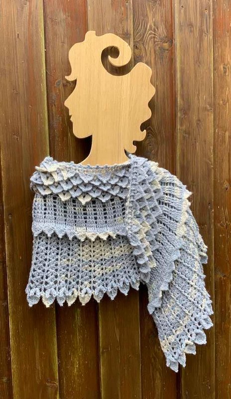 Hellblaues gehäkeltes Tuch mit Drachenschuppenrand, drapiert auf einer hölzernen Büste vor einer Holzwand.