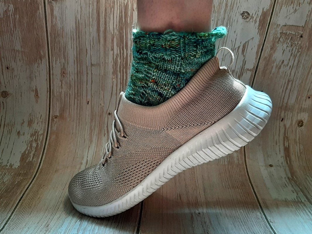 Handgestrickte grüne Socke mit Muster in beigem Sneaker auf Holzuntergrund.
