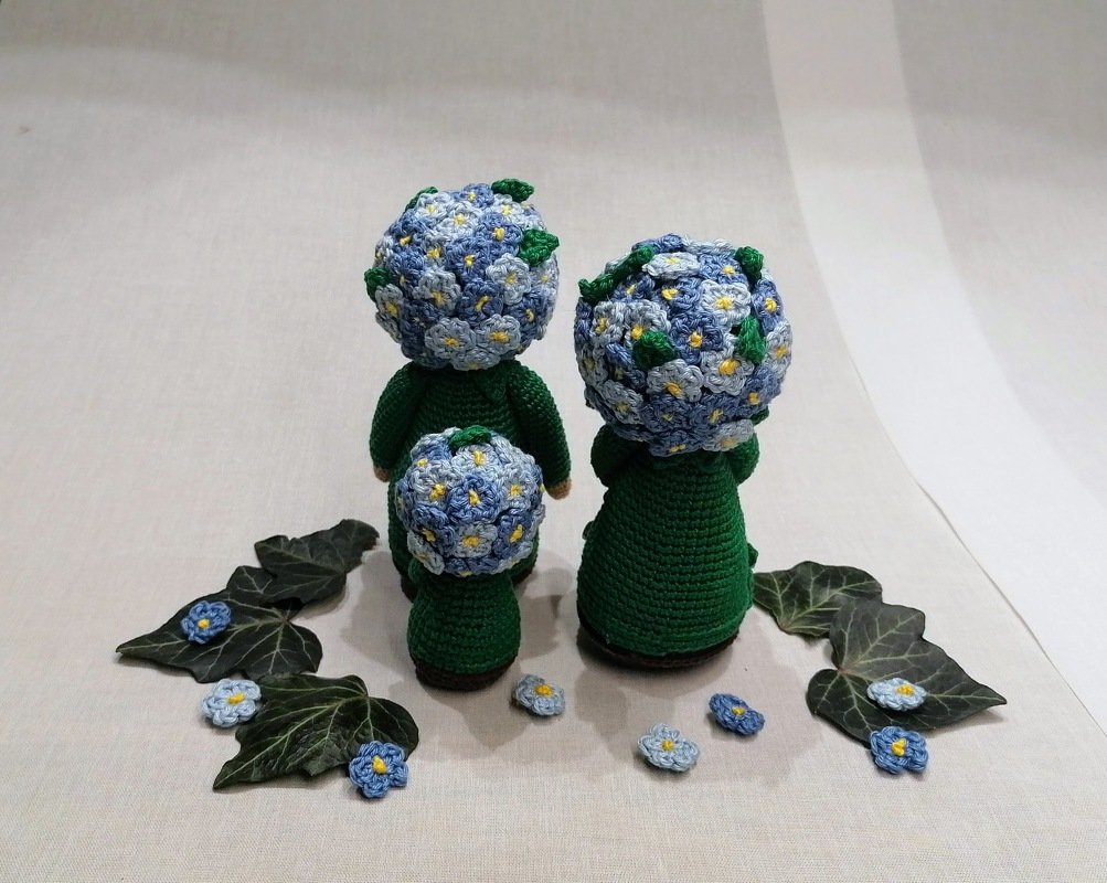 Drei gehäkelte Amigurumi-Puppen mit grünen Körpern und blauen Blütenköpfen, Rückansicht.