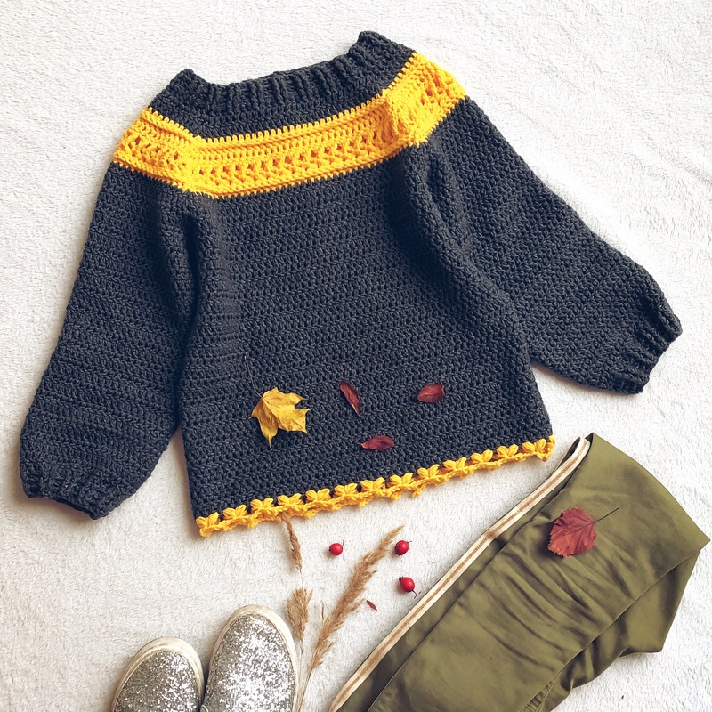 Dark gray crocheted women's sweater with a yellow chest stripe and yellow trim, styled with autumn leaves, red berries, glitter shoes, and olive pants.