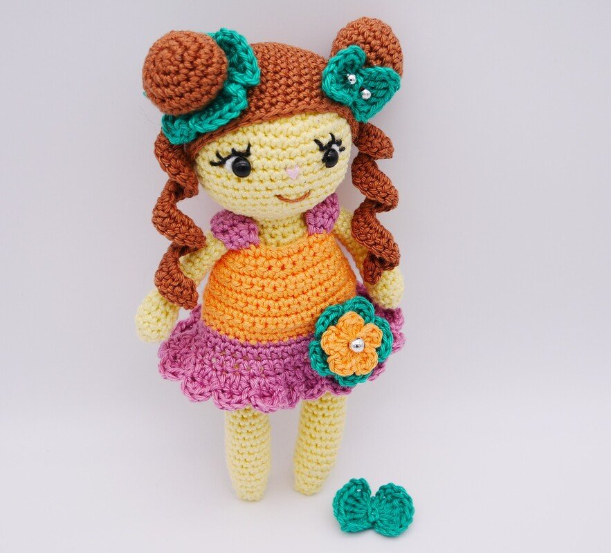Gehäkelte Amigurumi-Puppe mit braunen Zöpfen, orange-rosa Kleid und grüner Blumenapplikation