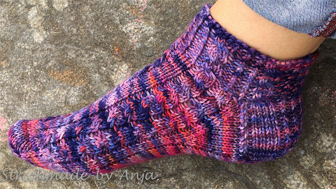 Gestrickte mehrfarbige Socke in Lila, Pink und Orange mit Zopfmuster an einem Fuß auf steiniger Oberfläche