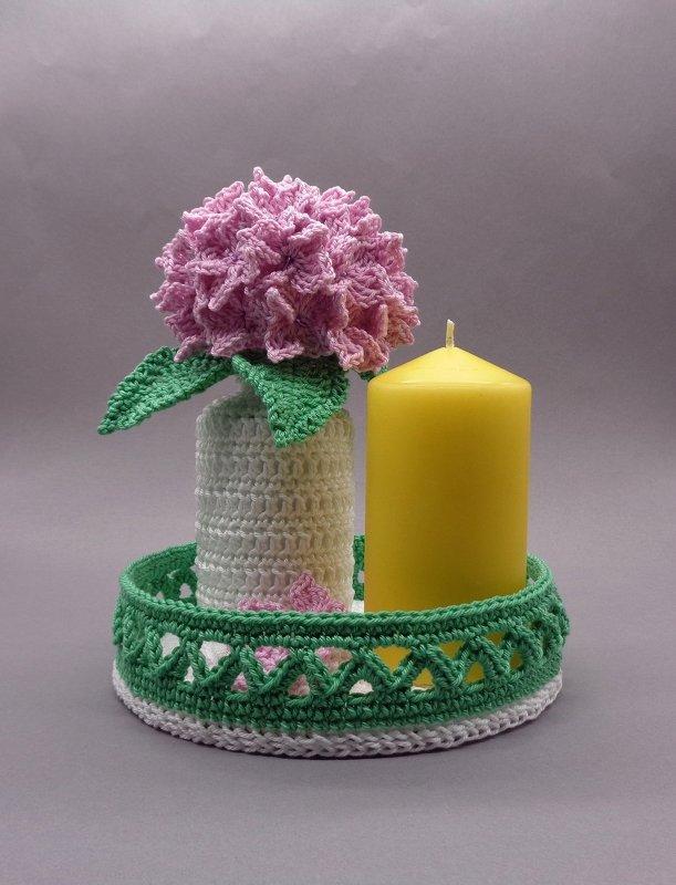 Rosa gehäkelte Blume in weißem Häkelgefäß neben gelber Kerze auf grün-weißem Häkeltablett