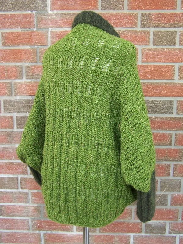 Rückansicht eines grünen, gestrickten Seelenwärmers mit vertikalen Lochmustern und dunklen Ärmeln vor einer Ziegelwand