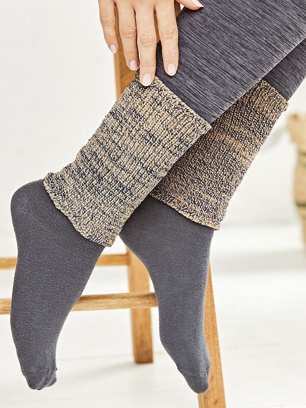 Beine mit melierten, beigen Strickbeinstulpen über grauen Leggings und Socken; Hand berührt die Stulpe.