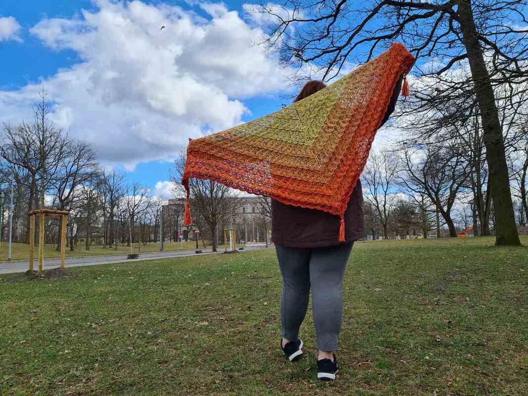 Person von hinten mit großem, dreieckigem Häkel­tuch in Gelb‑Orange‑Verlauf und Quasten in einem Park