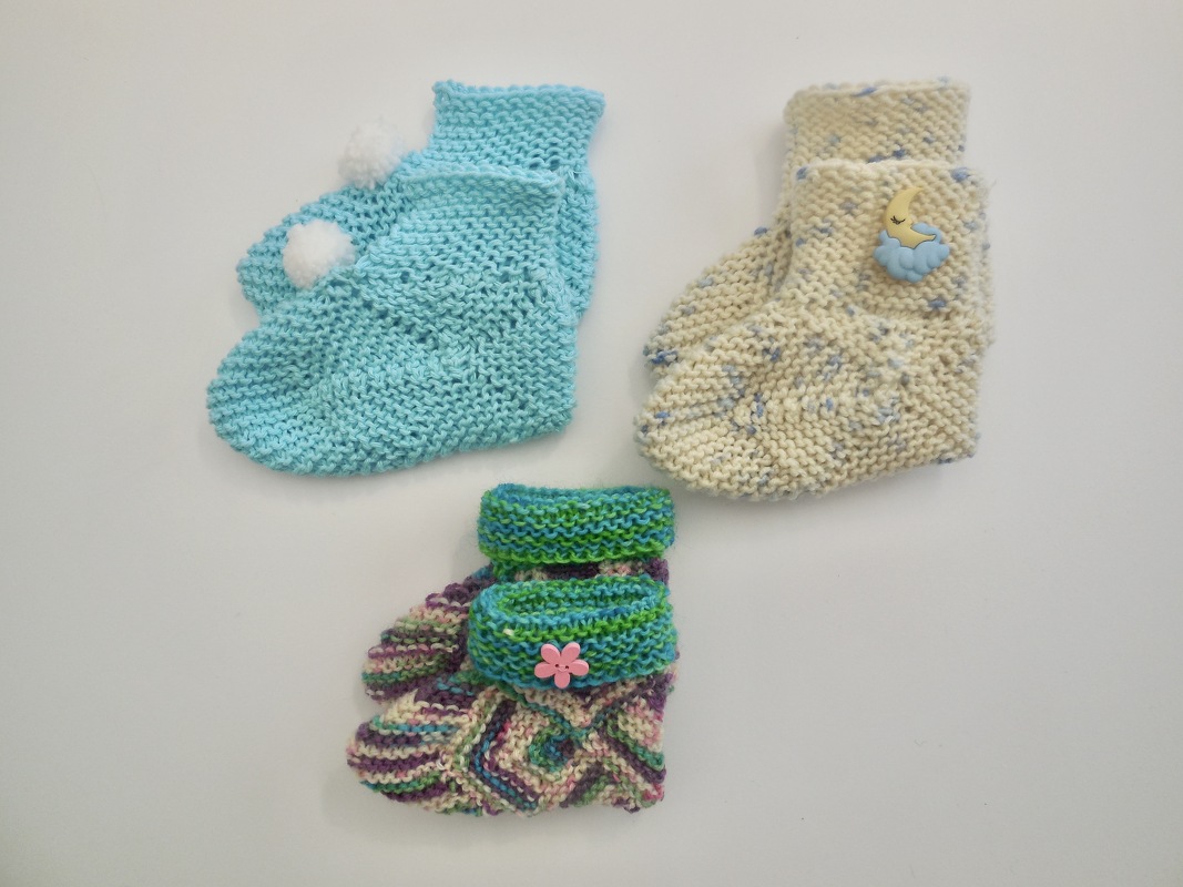 Drei Paar gestrickte Babyschuhe: türkis mit Pompons, cremefarben mit Mond- und Wolkenknopf, bunt gemustert mit Blumenknopf.