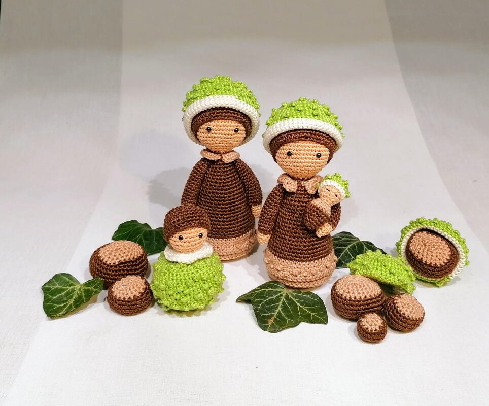 Gehäkelte Amigurumi-Kastanienfiguren: zwei große mit grünen Mützen, ein Baby, Kastanienschalen und Blätter.