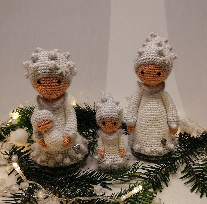 Drei cremefarbene Amigurumi-Puppen mit Noppenmützen auf Tannengrün und Lichterkette