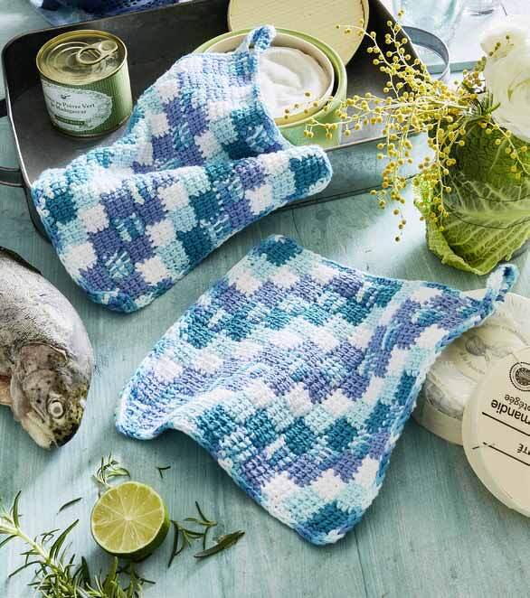 Zwei gehäkelte Topflappen in Blau und Weiß mit Karomuster auf Holztisch, umgeben von Fisch, Limette und Kräutern.