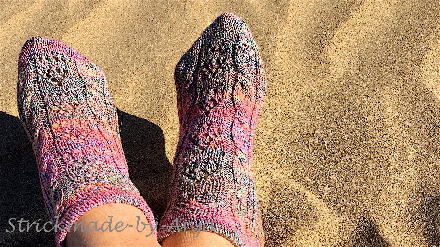 Ankle-length hand-knitted socks in variegated pink yarn showing lace stitch details on sand
