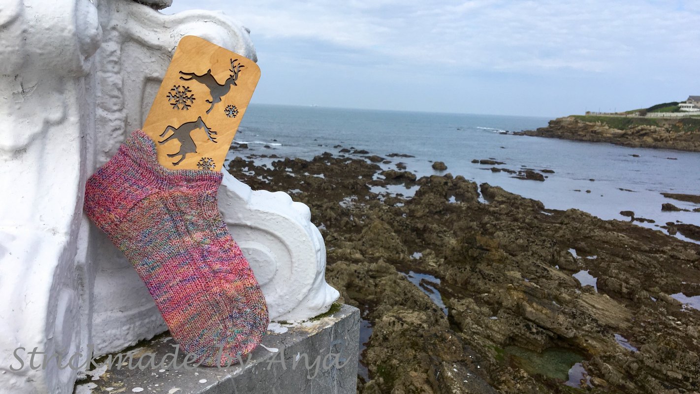 Multicolored hand-knitted ankle sock on a wooden sock blocker with deer and snowflake cutouts, set on a white ledge by a rocky coastline.