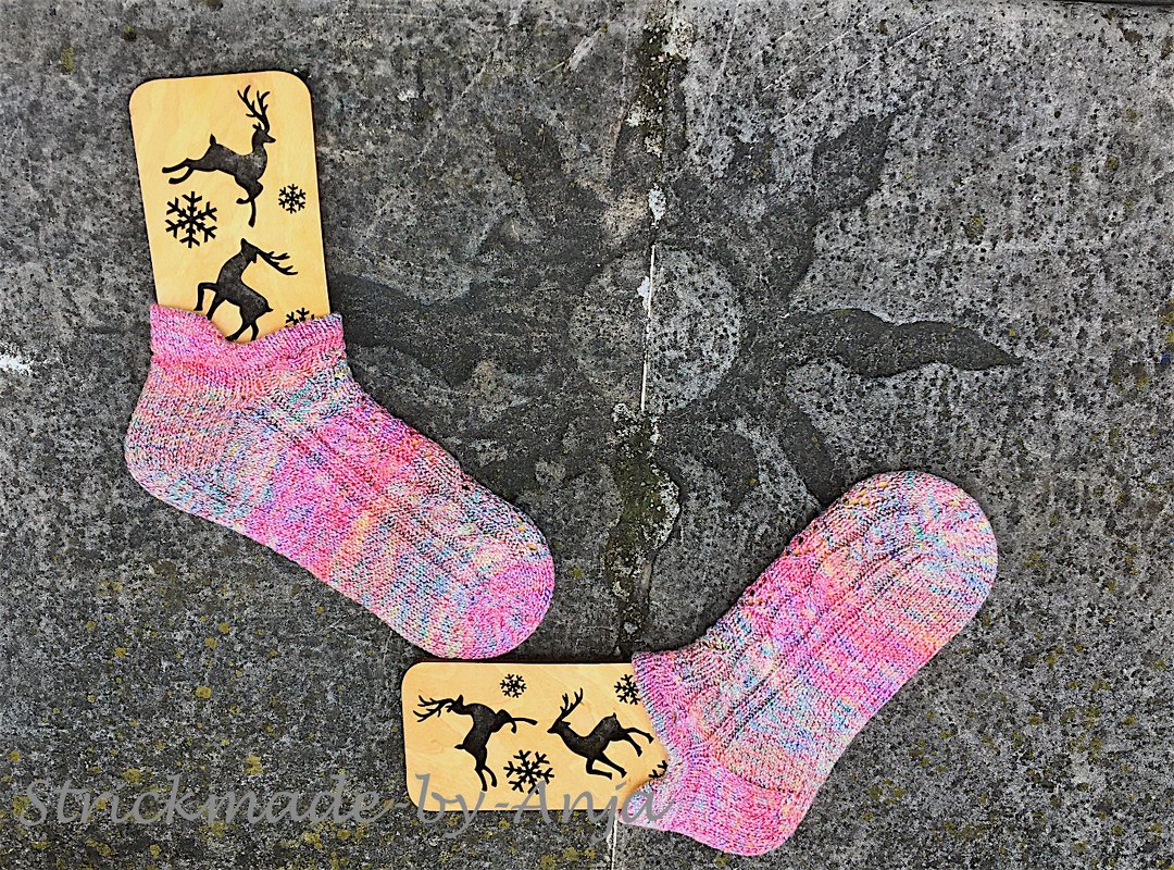 Pink and multicolored hand-knitted ankle socks on wooden sock blockers with deer and snowflake cutouts, on a gray textured stone surface.