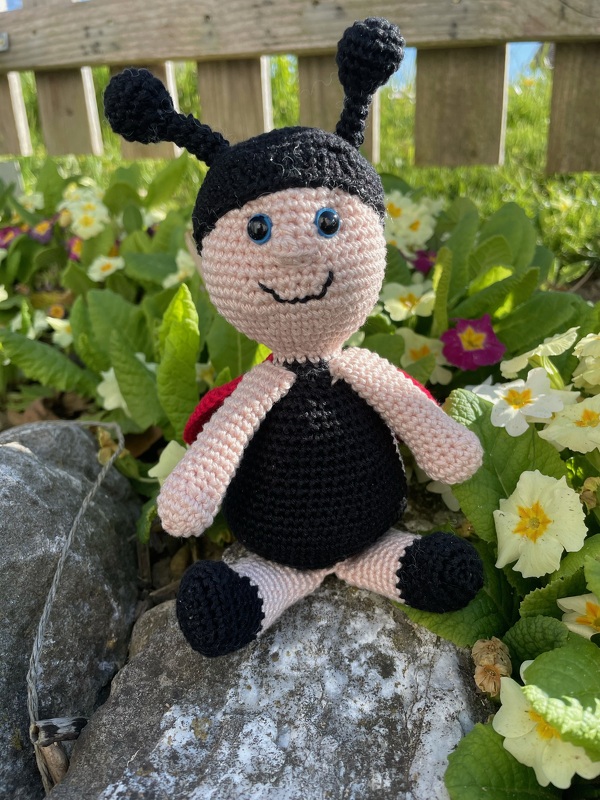 Handgehäkelter Marienkäfer-Amigurumi mit roten Flügeln und lächelndem Gesicht zwischen Frühlingsblüten