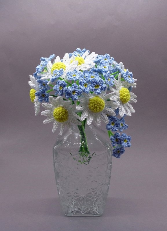 Gehäkelter Blumenstrauß mit weißen Margeriten und blauen Vergissmeinnicht in einer transparenten Glasvase vor neutralem Hintergrund.