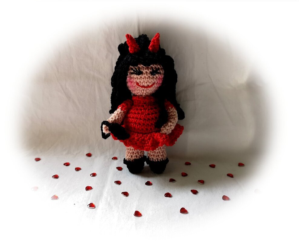 Gehäkelte Amigurumi‑Teufelspuppe mit roten Hörnern, schwarzem Haar und rotem Kleid vor hellem Hintergrund mit roten Herzchen.