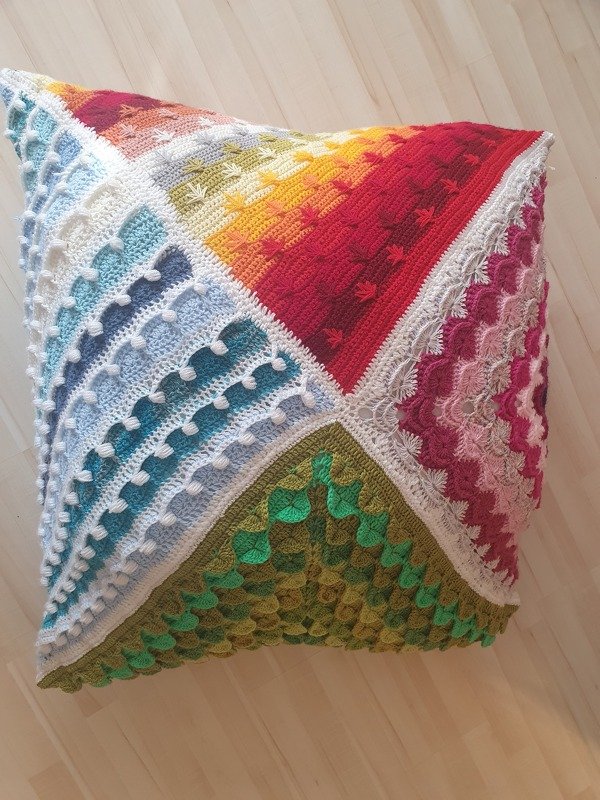 Quadratisches gehäkeltes Kissen mit vier bunten Paneelen, weißer Häkelkante und vielfältigen Reliefmustern