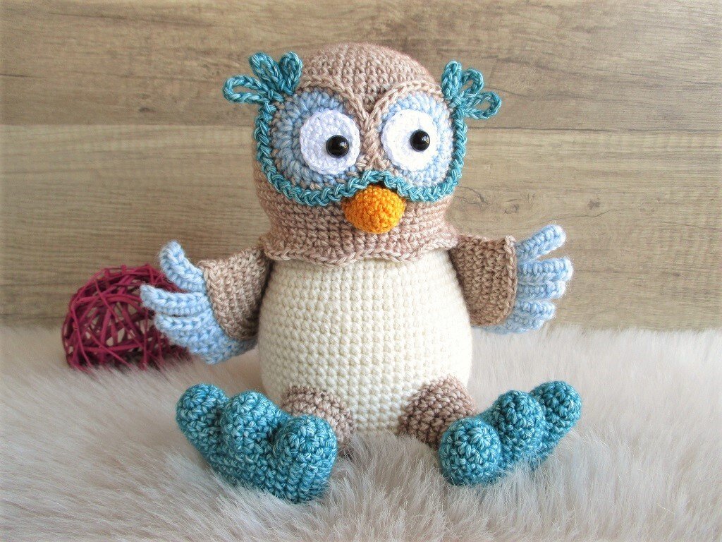 Sitzende gehäkelte Amigurumi-Eule in Braun, Creme und Blau auf flauschigem, hellem Untergrund vor einer Holzwand.