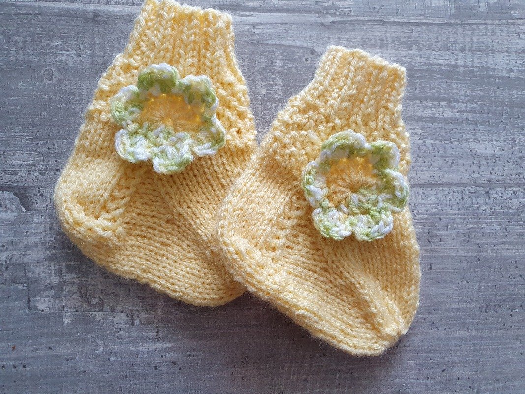 Zwei gelbe, handgestrickte Babyschuhe mit kleinen grün-weißen Häkelblumen auf grauer Holzfläche
