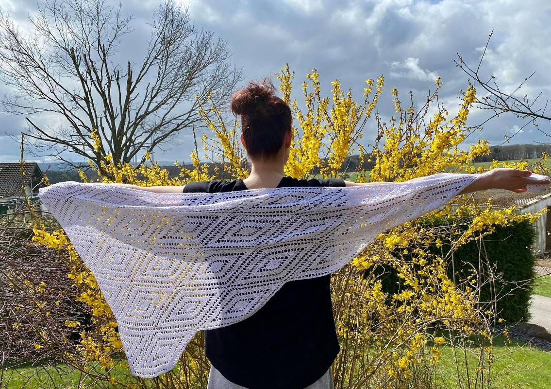 Person von hinten hält ein weißes gehäkeltes Dreieckstuch mit Rautenmuster vor gelb blühenden Sträuchern