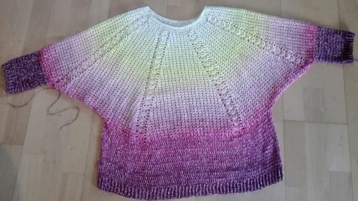 Gehäkeltes Oberteil mit weiten Raglanärmeln, Farbverlauf von hell zu dunkel und glitzerndem Garn, auf hellem Holzboden liegend.