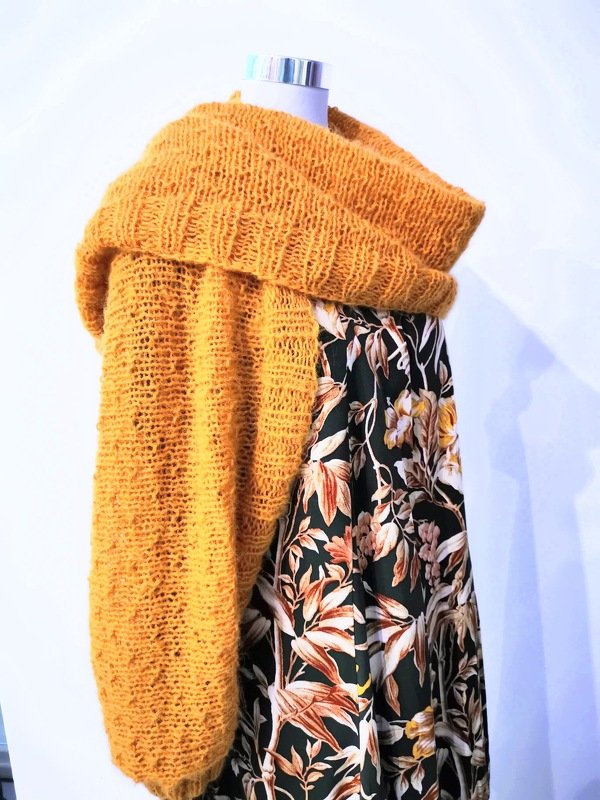 Senfgelber, locker gestrickter Shrug mit weiten Ärmeln über einem dunkelgrünen, geblümten Kleid auf einer Schaufensterpuppe.