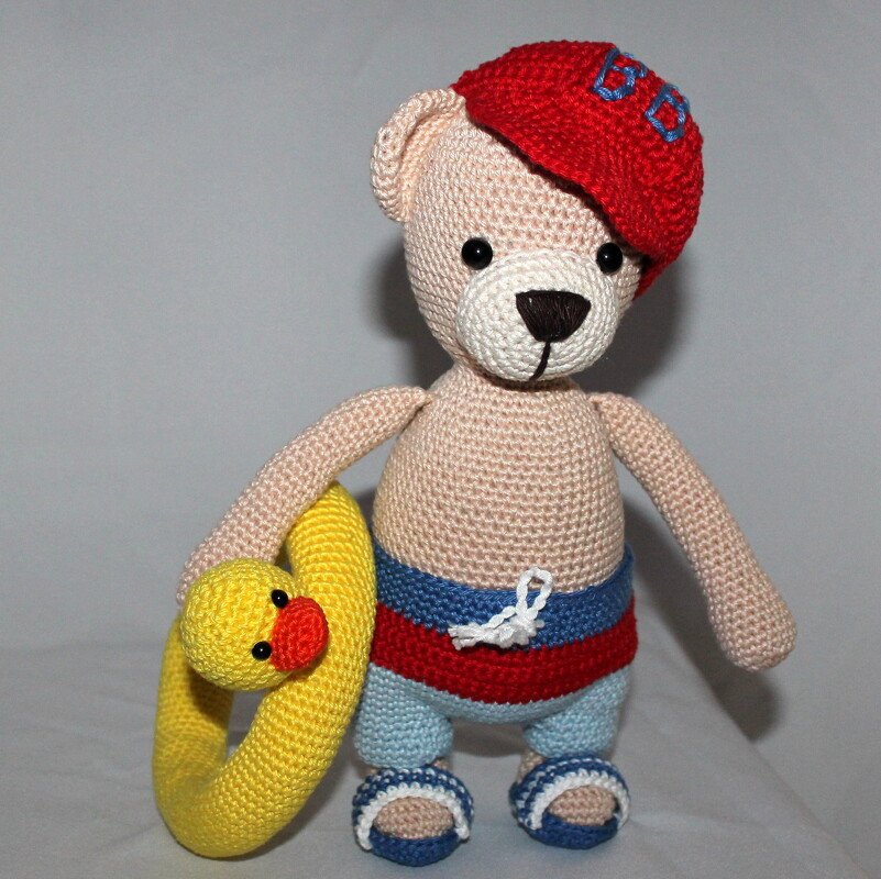 Beige amigurumi teddy in red cap holding yellow duck swim ring