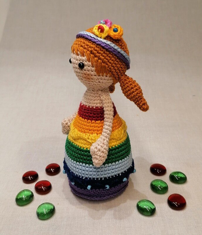 Gehäkelte Amigurumi-Puppe mit orangefarbenen Haaren, Blumenhaarband und gestreiftem Regenbogenkleid, davor rote und grüne Glassteine.