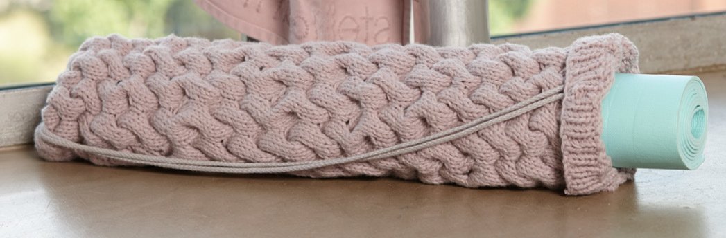 Gerollte mintgrüne Yogamatte in einer altrosafarbenen, grob gestrickten Hülle mit Zopfmuster und dünnem Kordelzug auf einem Fensterbrett.