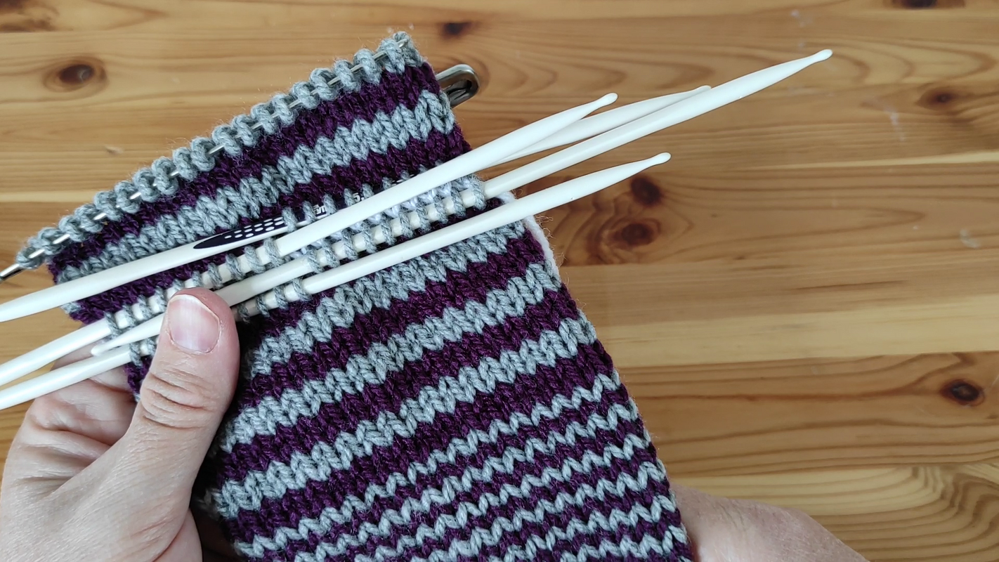 Hand hält rundgestricktes, gestreiftes Strickstück auf weißen Nadelspielnadeln