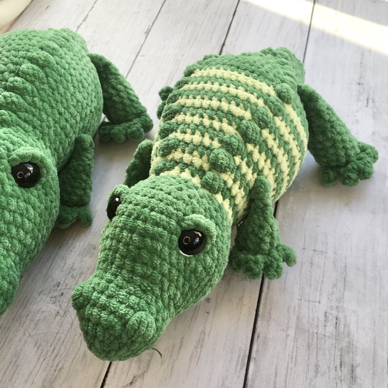 Two crocheted amigurumi alligators, one solid green and one striped green, on white wooden planks.