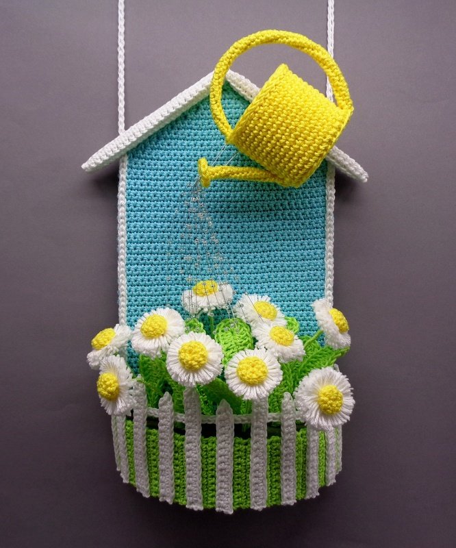 Crocheted house wall hanging with daisies, white picket fence and yellow watering can.