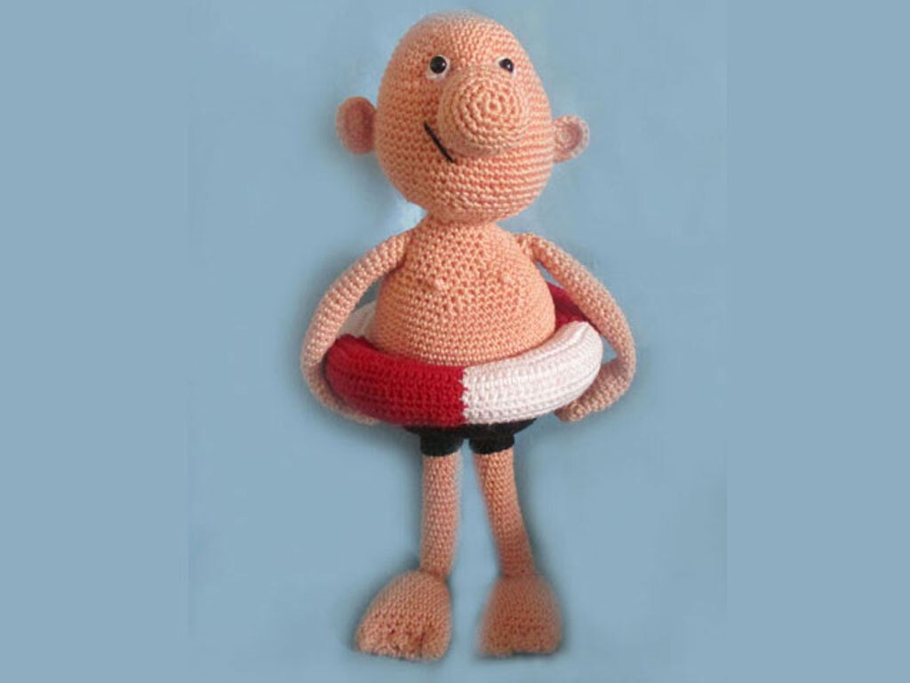 Gehäkelte Amigurumi-Puppe mit rosafarbenem Körper, schwarzer Badehose und rot-weißem Rettungsring vor blauem Hintergrund.
