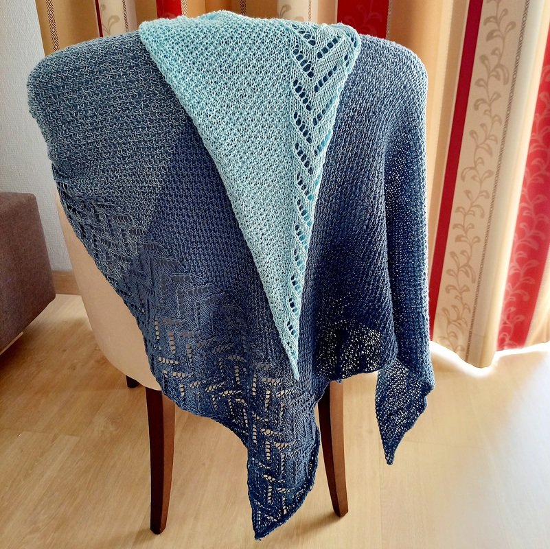 Two-tone blue ombre knitted triangle shawl with eyelet lace border and textured stitches draped over a chair.