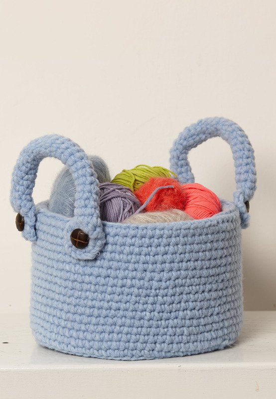 Hellblauer gehäkelter Korb mit zwei Griffen und braunen Knöpfen, gefüllt mit bunten Garnknäueln vor hellem Hintergrund.
