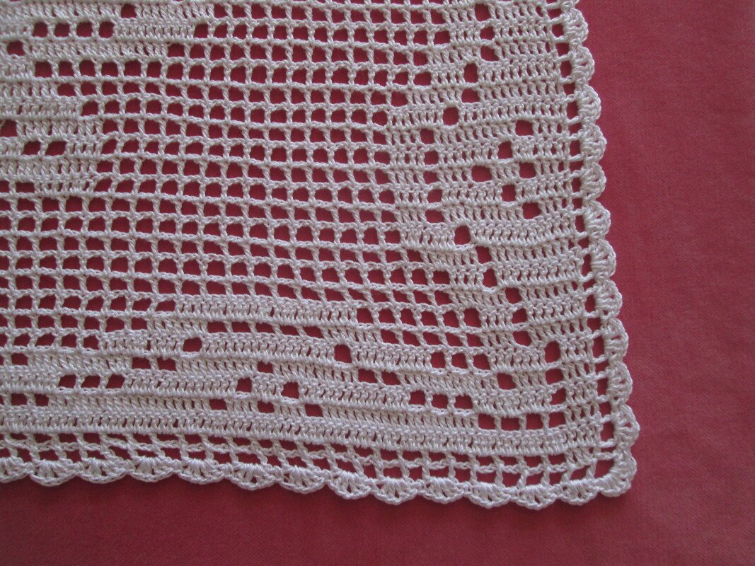 Weißes Häkeldeckchen mit Filetmuster und dekorativer Wellenkante auf rotem Hintergrund.