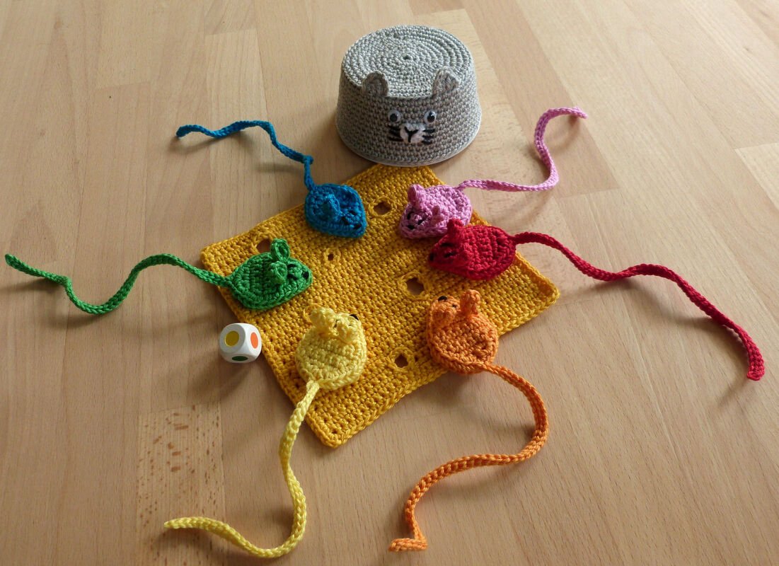 Sechs bunte gehäkelte Mäuse um ein gelbes Käsequadrat, dazu Farbwürfel und kätzchenförmiges Behältnis