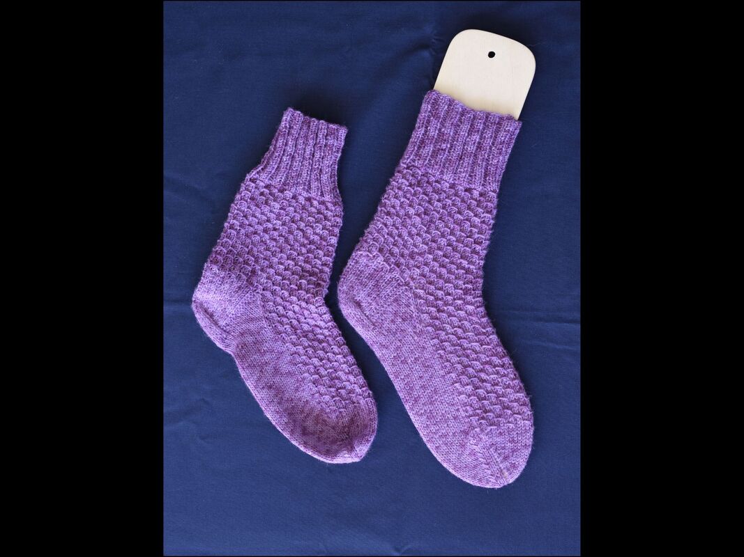 Zwei lila gestrickte Socken in unterschiedlichen Größen mit texturiertem Muster und Rippbündchen auf dunklem Stoff.