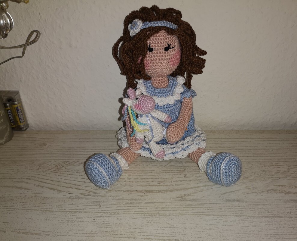 Sitzende gehäkelte Puppe mit braunen Locken, blau-weißem Kleid und kleinem buntem Einhorn im Arm.