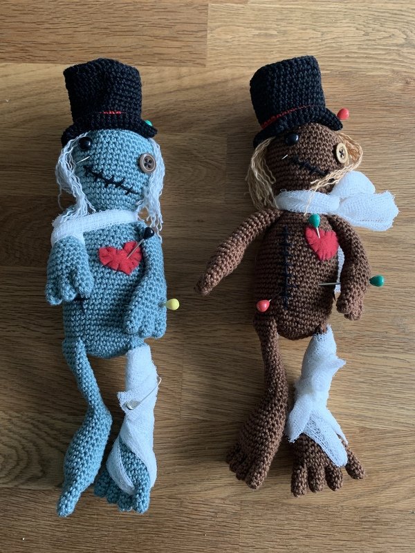Zwei handgehäkelte Amigurumi-Puppen mit Zylinderhüten, Filzherzen, Stecknadeln und Mullbinden auf Holz