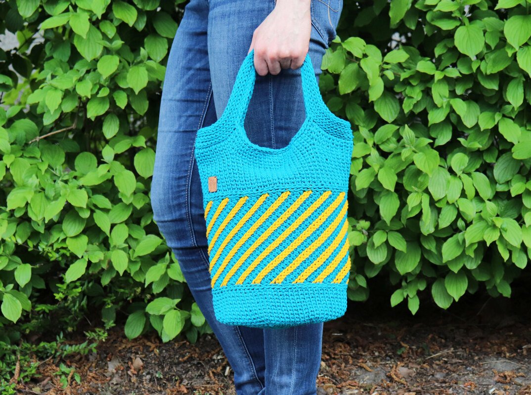 Gehäkelte Tasche in Türkis und Gelb mit diagonalen Streifen, an einem Griff gehalten.