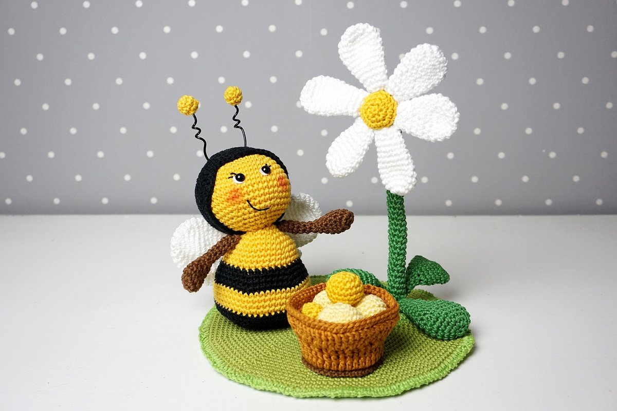 Gehäkelte gelb-schwarze Amigurumi-Biene mit weißen Flügeln neben gehäkelter Margerite und Körbchen