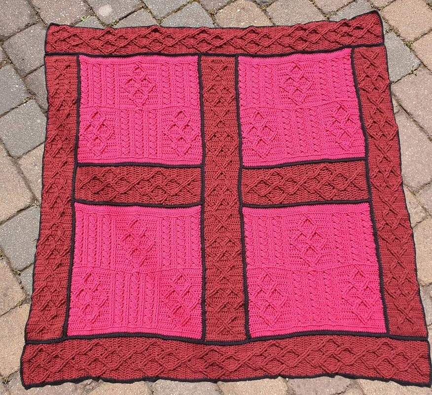 Gehäkelte Babydecke mit vier pinken Quadraten, bordeauxrotem Rand und schwarzen Nahtlinien, liegend auf Pflaster