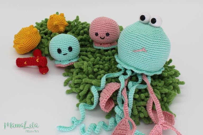 Gehäkelte Amigurumi-Meerestiere: große mintfarbene Qualle mit Augen, zwei kleine Kraken in Mint und Rosa, zwei gelbe Muscheln und ein roter Krebs auf grünem, zotteligem Stoff.