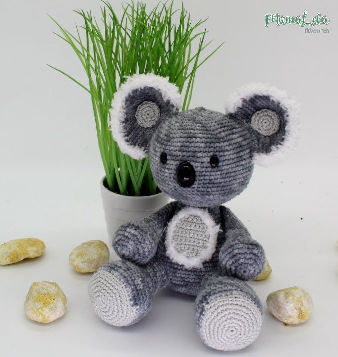 Gehäkelter Amigurumi-Koala in Grau und Weiß sitzt vor einer Topfpflanze.