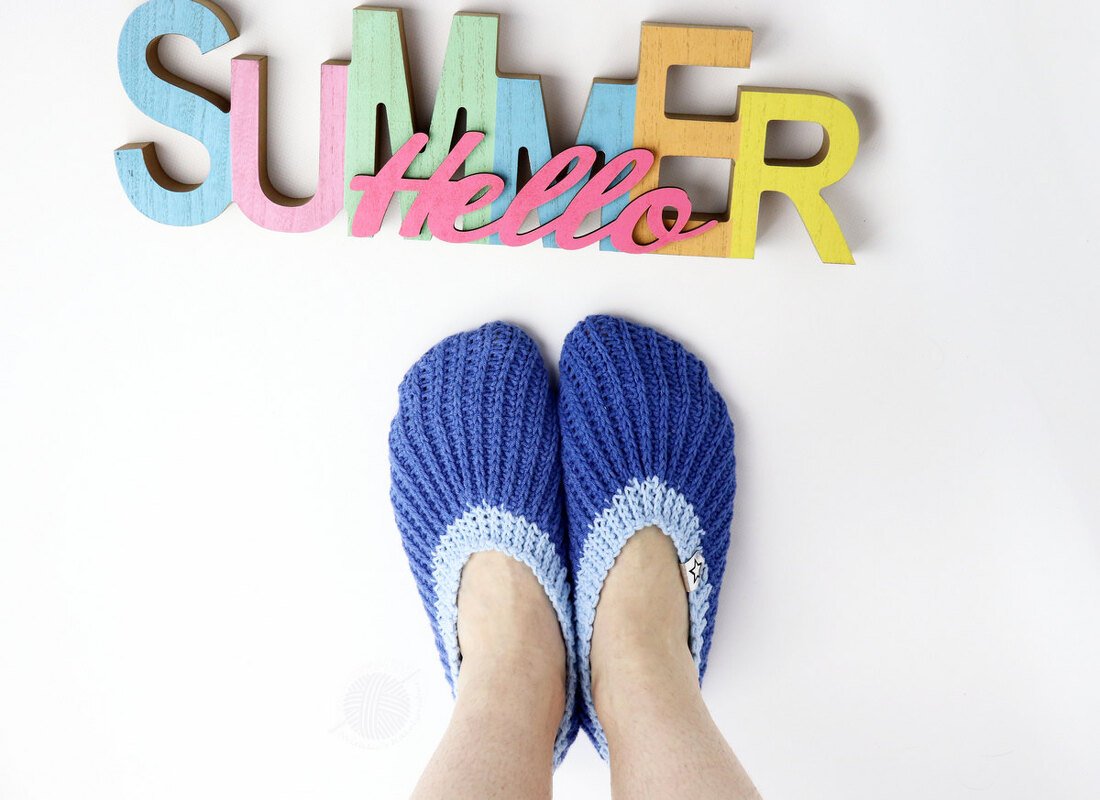 Blaue gehäkelte Pantoffeln mit hellblauem Bund vor bunten Holzbuchstaben 'Hello Summer'.